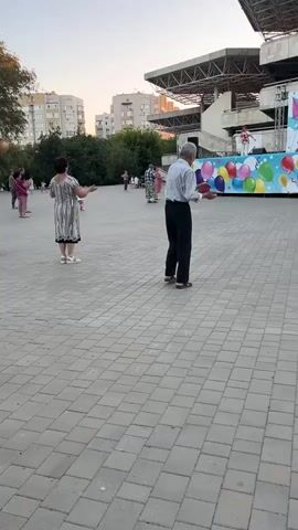 The video shows an outdoor scene at dusk. A group of people, primarily older adults, are gathered on a paved square. In the background, a stage is set up with a large banner featuring colorful balloons and text that appears to be a celebration. Several people are facing the stage, some with their arms raised as if participating in an activity or dance. A man and a woman in the foreground are facing away from the camera, also appearing to be engaged in some form of movement. The setting includes trees and apartment buildings in the distance, and the sky suggests it is either sunrise or sunset.