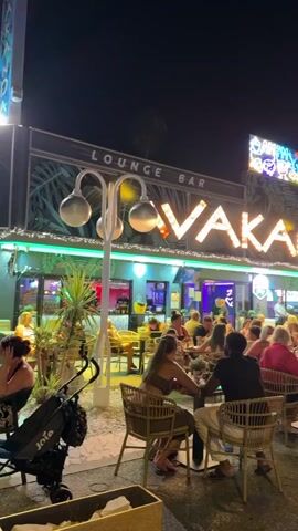 A nighttime scene on a street lined with palm trees. Several people are dining and socializing at tables outside a lounge bar named "VAKANDA." The bar has bright neon signage. People are walking along the sidewalk. A stroller is visible in the foreground.