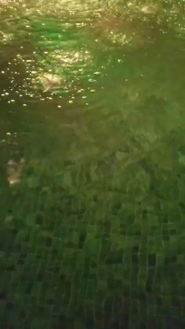The video shows a pool of water in what appears to be a dimly lit, artificial cave or grotto. The water has a greenish hue, possibly due to underwater lighting. Numerous bubbles rise to the surface. Thin streams of water fall from above, creating a curtain-like effect. In the background, a stone carving of a face is visible. A person is also partially visible in the background. The camera slowly pans and zooms in, providing a closer view of the details within the cave-like setting.