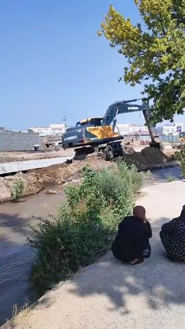 A Hyundai excavator is working on a construction site next to a body of water. Two people are sitting on the bank, watching the excavator. The excavator is digging a trench in the ground and moving dirt. The sky is clear and blue, and the sun is shining. There are trees and bushes around the construction site.