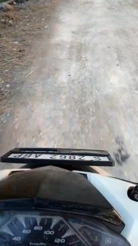 The video shows a first-person perspective of someone riding a white motorcycle on a dirt road. The camera is positioned above the handlebars, capturing the dashboard and the road ahead. The dashboard displays a speedometer with readings up to 140 km/h, but the needle is only slightly above 0, indicating low speed. A license plate is visible above the dashboard, displaying the characters "S 2862 ABP". The road is unpaved, with loose gravel and dirt. On the left side of the road, there are some dry plants and a wooden fence. The environment appears to be rural, with a sense of open space. The lighting suggests it is daytime, possibly late afternoon or early morning due to the soft light. The movement of the motorcycle is smooth, with slight bumps as it navigates the uneven terrain. The video captures the sensation of a leisurely ride through a natural landscape.