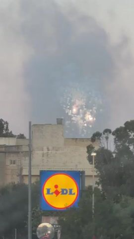 A drone shot from a low angle shows a hazy sky with smoke or fireworks obscuring the upper portion of the frame. Below the smoke, a building with a flat roof and light-colored walls is visible. A metal structure, possibly a chimney or ventilation unit, is situated on the roof. In the foreground, a large blue sign with the yellow and red Lidl logo is prominently displayed. The sign is mounted on a pole. To the right of the Lidl sign, a streetlamp illuminates the scene, and trees with dark green foliage are present. The lower part of the frame shows a rounded object, possibly a traffic mirror, reflecting lights, and the blurred outlines of a street. The video is shaky and slightly out of focus, especially at the beginning and end.