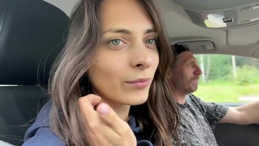 The video shows a young woman with long brown hair and green eyes in the passenger seat of a car. She is looking at the camera and smiling. A man with short brown hair and a beard is driving the car, visible in the background. The interior of the car is visible, including the dashboard and ceiling. The woman makes various facial expressions, including sticking out her tongue and laughing. The man in the driver's seat also appears to be speaking or reacting. The scene appears to be during daylight hours, with some greenery visible outside the car windows, suggesting they are driving through a rural or suburban area.