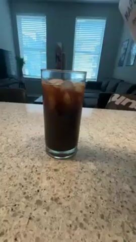 A hand pours liquid from a white carton labeled "The Maple Hill Difference" into a tall glass filled with ice and coffee. The glass sits on a speckled gray countertop in a kitchen with windows and a view of a living room. The carton is held over the glass, pouring a light-colored liquid, likely milk or a milk alternative, which swirls into the dark coffee. The scene is well-lit, with the countertop and background furniture in focus.