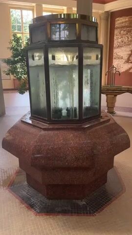 The video shows a close-up, stationary shot of an ornate fountain with a glass chamber filled with water. The fountain has a reddish-brown, speckled base and a hexagonal glass top section with brass trim. Inside the glass chamber, water is visibly circulating and bubbling, with some opaque material at the bottom. The fountain is situated in a well-lit, spacious room with a tiled floor, columns, and large windows in the background. A potted plant is visible to the left of the fountain, and a decorative fountain spigot is present on a tiered stand to the right. The overall impression is of an indoor public space, possibly a historical building or spa.