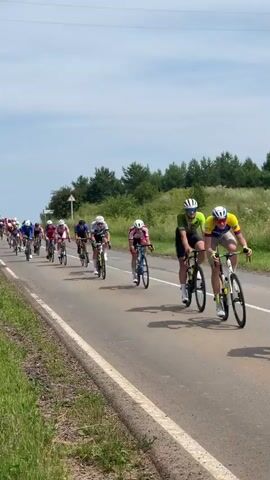 A group of cyclists in racing attire and helmets ride in a pack on a paved road. The road has a white line on the left edge and a grassy shoulder. Tall green trees and vegetation line the right side of the road, with a hazy sky above. The cyclists are positioned in a staggered formation, with one cyclist in the foreground on the right side of the road leaning forward over the handlebars. Another cyclist in a blue jersey is in the middle of the pack, also leaning forward. The cyclists are in motion, with the wheels of their bikes blurred. The sun appears to be shining brightly.