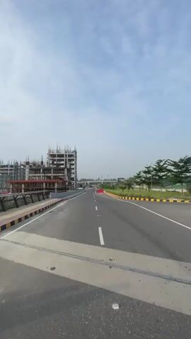 A first-person perspective video shows a person on a vehicle moving along an empty road. On the left side of the road, there is a large construction site with several unfinished buildings. On the right side of the road, there are trees and a median with yellow and black barriers. The road is clear of other vehicles, and the sky is partly cloudy. The video appears to have been recorded during the daytime.