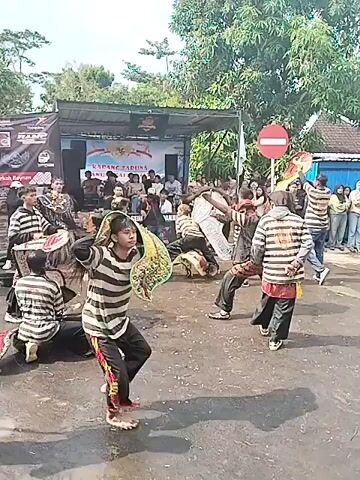 A group of people are performing a traditional dance in an outdoor setting. Many of the dancers are wearing black and white striped shirts with black pants. They are also holding decorative masks that resemble the heads of mythical creatures. The masks are brightly colored. The dancers are moving in a coordinated manner. In the background, there is a stage with people sitting on it. There is a crowd of people watching the performance.  There is also a "do not enter" sign visible near the stage. The dance is taking place on a paved surface. There are trees and buildings in the background. The overall atmosphere seems lively and celebratory.