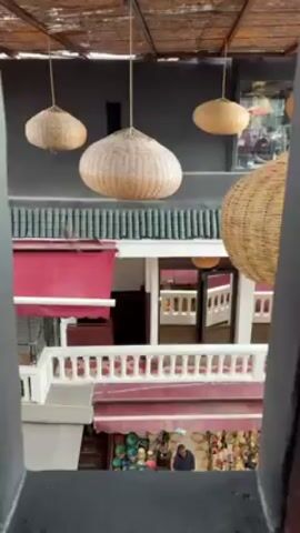 A high-angle view from a window shows an outdoor market. Several large, woven, spherical light fixtures hang from the ceiling above. The scene looks down into a multi-level courtyard of shops. The buildings are painted in shades of white, grey, and red. Tourists are browsing the merchandise displayed outside the shops, including hats and various souvenirs. The camera slowly pans to reveal more of the marketplace below.