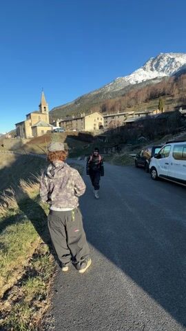 The video shows two individuals standing on a road in a mountainous, rural setting. One person, wearing a camouflage hoodie, baggy pants, and a hat, stands near the edge of the road, facing away from the camera. The other person, dressed in a black puffer jacket, dark pants, and sunglasses, is standing on the road facing the first person. They reach out and do a fist bump. In the background, a traditional European village with stone buildings and a church with a tall steeple is visible. A snow-covered mountain rises in the distance under a clear blue sky. A white van with the words "La Region" and "A-PIN" written on it is parked along the side of the road.