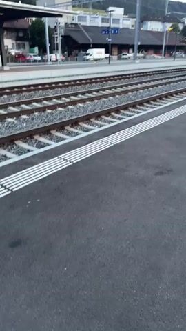 The video shows a train platform with multiple railway tracks. On the right side of the frame, a white and red train is speeding past the platform. The train has several windows, some of which reflect the surrounding environment. The platform has a textured surface with white tactile paving strips along the edge. In the background, there are buildings, a street with cars, and some greenery on a hillside. The sky is visible in the upper part of the frame.