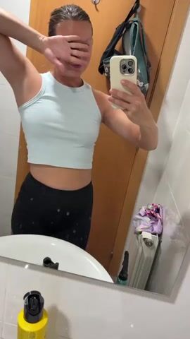 A woman stands in front of a mirror, taking a selfie. She is wearing a light blue crop top and black leggings. Her left arm is raised, and she is covering her eyes with her hand. In the mirror's reflection, a backpack hangs on a door, and a radiator with laundry draped over it is visible. The room appears to be a bathroom.