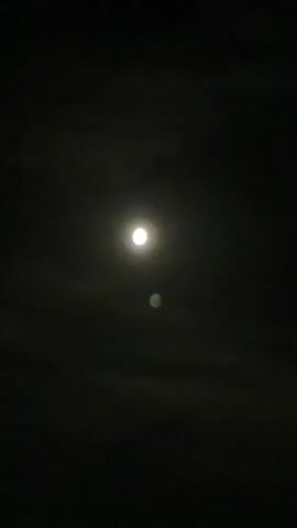This is a short video of the moon in the night sky. The moon is bright and appears to be full. There are some clouds in the sky, which partially obscure the moon at times. There is also a faint green glow below the moon, which may be lens flare or another celestial object. The video is dark overall, with the moon being the only significant light source.