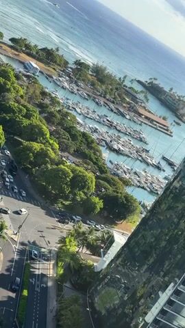 A high-angle shot shows a marina filled with sailboats and yachts. The water is a deep blue, transitioning to a lighter turquoise closer to the shore. Several boats are docked in neat rows. Beyond the marina, there is a coastline with trees and some buildings visible. In the foreground, there is a street with cars parked and driving. Lush green trees are abundant in the middle ground, bordering the marina and the street. The sky is partly cloudy with bright white clouds against a clear blue sky. The overall scene depicts a sunny day at a coastal location.