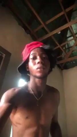 A shirtless dark-skinned man with a goatee and a red and black baseball cap backwards is in a room with a wooden ceiling. He is smiling and looking at the camera. The video is taken from a low angle. The man has a gold chain around his neck. There is a doorway visible in the background.
