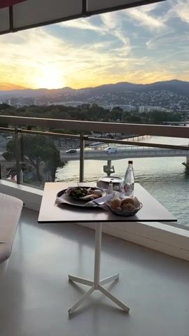 The video shows a table set for a meal on a balcony overlooking a scenic view. The table is square and white, supported by a white pedestal base. On the table, there is a black tray with a plate of food featuring a salad and what appears to be a cheese disc, a cloche lid over another dish, silverware wrapped in a white napkin, a salt and pepper shaker, a bottle of V water, a drinking glass, and a basket of bread rolls. The background reveals a sunset with a mix of yellow and orange hues, silhouetting a mountainous cityscape. Below, there is a body of water with a bridge, and some greenery on the shores. The balcony has a glass railing with a silver metal frame. The flooring of the balcony is light grey. The video slowly zooms in on the table setting.