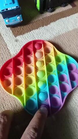 The video shows a hand interacting with a rainbow-colored pop-it toy. The pop-it is in a cloud-like shape and has multiple rows of bubbles. The hand presses down on the bubbles, causing them to pop. In the background, two toy vehicles are visible: a blue bus with a face and a green and black truck. The objects are on a textured surface that appears to be a rug or carpet.