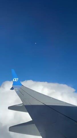 The video shows an airplane wing against a backdrop of blue sky and clouds. The wing, painted in shades of gray, bears a blue logo on its tip. The clouds form a layer beneath the wing, while the clear blue sky stretches above. In the distance, a faint, circular object is visible in the sky, possibly the moon. The video is taken from inside the airplane, offering a passenger's perspective.