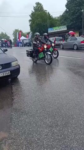A person rides a vintage motorcycle on a wet road during what appears to be a car show or parade. It is raining, and several cars are parked along the side of the road. Spectators are visible holding umbrellas and observing the event. The motorcycle is orange and black with saddlebags on the back. The rider is wearing a helmet and a jacket. A white pickup truck is visible in the background, as are several other vehicles and tents.