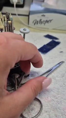 The media file shows a close-up of a person's hands interacting with dental or jewelry tools. The scene appears to be indoors, on a white, textured surface, possibly a towel or cloth. In the background, a rack holds various small, cone-shaped and disc-shaped burrs or polishing tools, commonly used in dental work or fine crafting. A box labeled "Whisper" is also visible. The lighting suggests an indoor setting, with no indication of time of day or weather. The person's hands, one with a manicured nail, are manipulating a pair of small, precision scissors and a metal instrument with a fine point. The activity suggests meticulous work, perhaps preparing or cleaning a tool, or positioning a small object. There is a blue sticky note with some handwritten text in the background, but it is illegible. There are also small, dark blue rectangular pieces of material on the white surface, the purpose of which is unclear. The overall impression is of a detail-oriented task being performed in a workshop or laboratory setting.