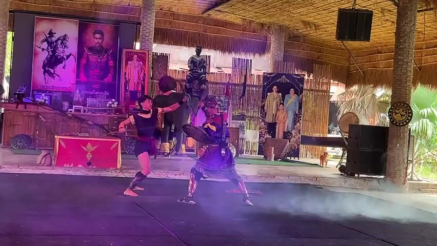 Two individuals are engaged in a mock sword fight on a stage. The person on the left wears a dark vest and shorts with arm and leg wraps, wielding a sword. The person on the right is dressed in purple and gold armor, also holding a sword. They are positioned under a thatched roof structure. The stage is dimly lit with purple lighting on the left side. In the background, there are several large posters depicting historical figures and what appear to be royal portraits. There is also a large, dark figure that resembles an elephant statue. Smoke or mist is present on the stage, obscuring the lower portions of the fighters and creating a dramatic effect. The overall environment suggests an outdoor performance space, possibly a tourist attraction or cultural center.