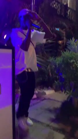 A person wearing a white t-shirt and a baseball cap is holding a microphone to their mouth, appearing to be singing or speaking. They are wearing a cross necklace, a bracelet on their left wrist, and a crossbody bag. The background is dark, with purple and white lights creating an atmospheric effect. There are some palm leaves and other foliage visible, suggesting an outdoor or semi-outdoor setting. The person is gesturing with their right hand.