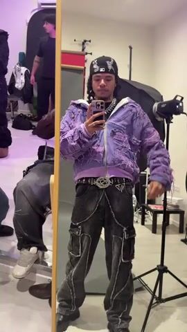 A young man takes a selfie in front of a mirror, showing off his outfit. He wears a black bandana, a purple hooded jacket with frayed details, black cargo pants, and a distinctive belt. The environment appears to be a studio or dressing room with lighting equipment visible in the background. Several other people can be seen in the reflection and periphery.
