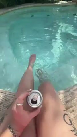 A person sits by a round pool, dipping their feet in the water. They are holding an open aluminum can in their hand. The pool water is clear and light blue. The pool is surrounded by light brown tiling. The person has pale skin with tattoos on their legs. They are wearing a ring on their finger. The background shows green foliage.