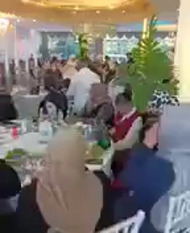 This video shows a crowded indoor event, possibly a banquet or celebration, with many people seated at tables. The environment appears to be a large hall with a decorative, circular ceiling fixture that is illuminated. There are also plants visible, and the lighting suggests an evening or indoor event. The people in the video are dressed in various attire, and some appear to be interacting with each other. The camera is moving, providing a panoramic view of the event.