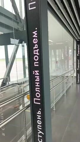 The video appears to be taken inside an airport terminal, likely at Saburovka, Russia, given the textual context. The scene is a bright, modern airport walkway with large glass windows overlooking an airfield. The architecture features exposed metal beams and a slatted ceiling.

The primary subjects are a woman carrying a child on her back, and a person walking in the distance. The woman is wearing a backpack and carrying a large black bag. She is walking away from the camera down the corridor. There are also escalators visible in the background, suggesting movement within the terminal.

The activity depicted is movement through the airport, likely towards a gate or exit. The overall impression is one of transit and travel. The lighting suggests it is daytime, and the cloudy sky seen through the windows indicates overcast weather.

Several pieces of text are visible on digital displays. On a vertical pillar, text in Russian reads "Полный подъем" (Full ascent). On horizontal displays and vertical pillars further down the corridor, the word "Поехали!" (Let's go!) is repeatedly displayed in Russian. This phrase is a common exclamation for starting a journey. The presence of these messages, particularly "Let's go!", reinforces the theme of travel and departure.