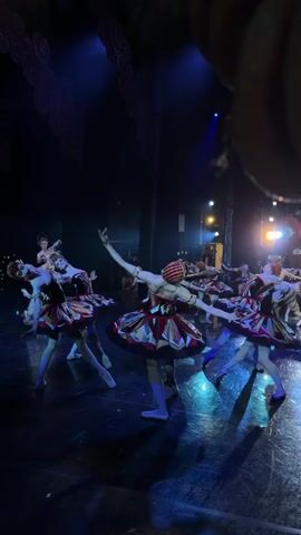 This media file captures a ballet performance on a stage. The scene is dimly lit with theatrical blue and white spotlights, creating a dramatic atmosphere. The stage floor reflects the lighting, adding to the visual spectacle.

The primary subjects are a group of female ballet dancers. They are dressed in elaborate costumes featuring short, layered tutus in red, gold, and black, with intricate detailing. Each dancer also wears a matching headpiece adorned with gold and red elements, resembling a crown or elaborate headdress. Their attire suggests a performance of a fantastical or historical nature, possibly from a ballet like "The Nutcracker" given the festive costumes.

The dancers are engaged in a coordinated ballet routine. Their poses are graceful and expressive, with arms extended and bodies arched, demonstrating classical ballet technique. Their faces, though not perfectly clear due to the lighting and distance, convey focused concentration and artistic expression. The dancers are arranged in a circular formation, moving in unison.

The background of the stage is dark, with some hints of theatrical backdrops or stage machinery visible, further emphasizing the focus on the performers. The overall mood is one of elegance, artistry, and theatrical magic. There is no visible text in the media. The context provided indicates this performance is taking place in Ufa, Russia.