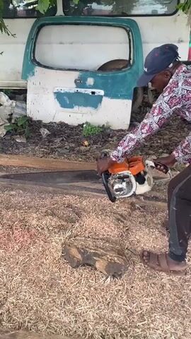 An adult male, barefoot and wearing a dark baseball cap and a long-sleeved, patterned shirt, is operating an orange and black STIHL chainsaw. He is crouched, guiding the saw's long bar through a piece of timber laid horizontally on the ground. A substantial amount of light-colored sawdust and wood shavings covers the ground around the cutting area, with a smaller, rough-hewn log visible in the foreground. In the background, a large, painted metal panel, appearing to be the rear door or tailgate of a vehicle (white with a light blue lower section), leans against an unseen structure. Sparse green foliage and dry leaves are visible on the ground behind the panel. The scene depicts an outdoor woodworking activity under bright daylight, suggesting a rustic or workshop setting. The man appears focused on his task.