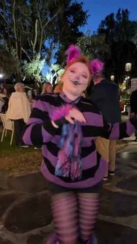 A young woman, dressed as the Cheshire Cat, is featured in a dynamic video shot outdoors at night in Nir Eliyahu, Israel. She wears a distinctive costume consisting of a purple and black striped sweater, black shorts, and matching striped tights. Her accessories include a pink fur cat-ear headband, pink fur wrist cuffs, and black fingerless gloves with pink paw print details. A purple and blue patterned scarf is tied around her neck. Her face is elaborately painted with a purple nose, white fangs, and black whiskers, accentuated by vibrant pink eye makeup.

Throughout the video, she poses playfully, making cat-like gestures with her hands and displaying energetic, mischievous facial expressions. She smiles widely, showcasing her painted fangs, and at one point, leans in close to the camera with a grin. In the background, other people can be seen at what appears to be an evening outdoor gathering or party. The scene is illuminated by string lights strung between trees, indicating the nighttime setting. The atmosphere is lively and festive, reflecting the character's whimsical nature.