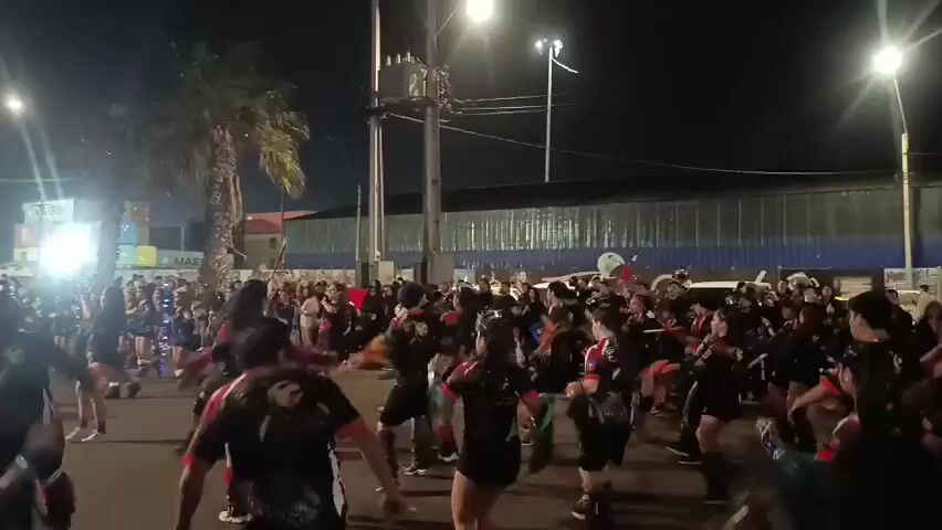 A large group of people are dancing and performing in a street at night. They are dressed in matching black and red outfits, and many are holding sticks or poles that they are swinging around. There are streetlights and palm trees visible in the background. The atmosphere appears to be festive and energetic.