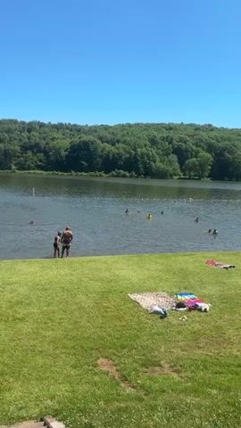 A full, vertical, outdoor shot shows a lake with many people swimming and relaxing on the shore on a clear day. The lake is lined with trees and a few people in the water. The shore has green grass with several blankets laid out for sunbathing and relaxing.
