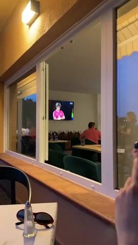 The video shows a view through a window of people sitting in what appears to be a cafe or restaurant. A television screen is visible on the wall, displaying a soccer match. A young man in a pink jersey is shown holding a soccer ball. Another man in a suit and tie is also on the screen. Inside the cafe, a man in a red shirt is seated at a table, facing away from the camera, looking at the TV. The cafe has green upholstered chairs and wooden tables. Outside the window, a person's hand holding a blue object is visible in the lower right corner. The lighting suggests it is evening.