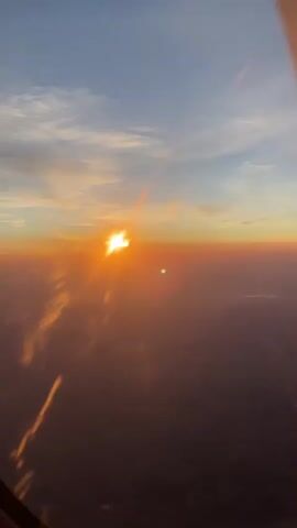 The video shows a view from an airplane window during a sunset. The sun is low on the horizon, casting a warm orange glow across the sky. The clouds are illuminated by the setting sun, and some appear to be reflecting the light. Below, the earth is visible as a dark, shadowed landscape. The wing of the airplane is visible in the upper right corner of the frame.