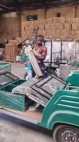 A muscular man wearing a tank top and dark pants is standing in a large warehouse filled with boxes, loading what appears to be a rolled-up canvas or tarp into a green three-wheeled electric vehicle. The vehicle is open in the back, and already contains several window-like panels wrapped in plastic. Another similar vehicle is visible behind the man.  A ladder leans against a stack of boxes in the background.