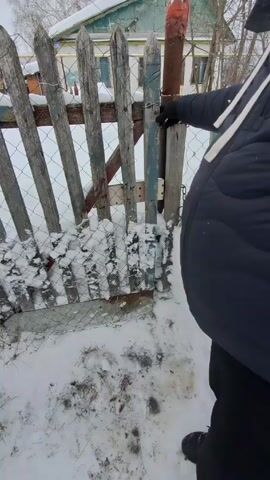 Here's a description of the video:

A person wearing a dark winter coat, gloves, and dark pants is standing in the snow next to a weathered, wooden gate covered in snow. The gate is held shut by a piece of twisted wire. The person reaches out, untwists the wire, and opens the gate. Beyond the gate is a snow-covered area, and in the background, a small house is visible with snow piled around it. The environment is clearly winter with a thick layer of snow on the ground and structures.