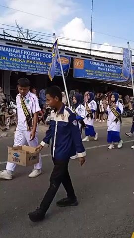A group of adolescents in matching parade outfits are walking down a street. They wear dark blue and white uniforms, some with white lab coats and hijabs. Several carry flags with crests. They wear sashes that identify them as members of an organization. Shops are lined along the street, with signage advertising frozen foods and plastic shop services. Spectators watch from the sidewalks. A young girl in a yellow dress stands in the foreground. The sky is bright with scattered clouds.
