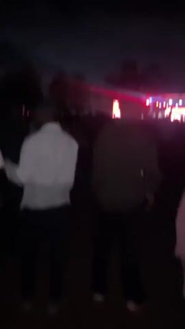 The video shows a crowd of people at an outdoor event at night. There is a stage with bright lights in the background, suggesting a concert or performance. The people are moving and dancing. Some individuals are wearing light-colored shirts, while others are in darker clothing. One person wears a cap. The atmosphere appears lively and energetic.