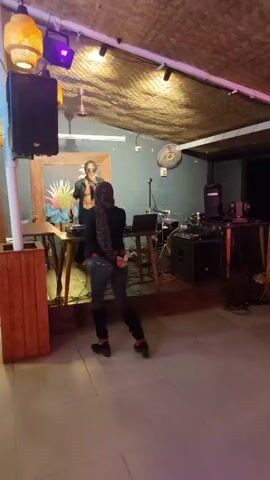 The video shows an indoor scene, likely a bar or restaurant, with a thatched roof and rustic decor. The lighting is dim, with a mix of natural light filtering through openings and artificial colored lights, suggesting it's evening or nighttime. A DJ setup with speakers and equipment is visible, indicating a musical performance or party is taking place.

In the foreground, a person in dark clothing with a headscarf is facing away from the camera, walking towards a stage area. On the stage, a performer, possibly a singer or rapper, is holding a microphone and appears to be interacting with the audience. The performer is wearing sunglasses and a suit jacket.

Further back, a person with long, braided blonde hair is holding up a smartphone, seemingly recording or taking pictures of the event. Other individuals can be seen in the background, some seated at tables. Red and blue laser lights are projected onto the floor, adding to the festive atmosphere. The overall mood seems lively and engaging, consistent with a performance or social gathering. The mention of "Mandrem, India" as the location context suggests a tropical or subtropical setting, which could influence the architectural style and the general ambiance of the venue.