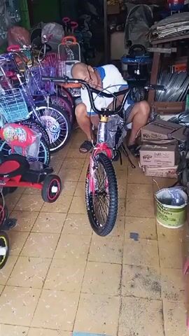 The video shows a person asleep on a red bicycle inside a store. The person is wearing a blue shirt and shorts and is resting their head on the handlebars. The bicycle is a children's bike with training wheels. There are other bicycles and tricycles in the background, as well as shelves stocked with various items. The floor is tiled and the lighting is dim.