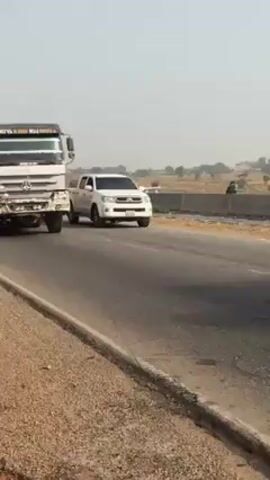 The video shows a road in an arid landscape under a clear sky. A large truck passes the camera’s position on the roadside, moving from right to left, followed by a white pickup truck. As the camera pans to continue following the road, a yellow auto-rickshaw, or tuk-tuk, is seen approaching from the distance and also passes the camera position. The road appears to be a major highway, with a shoulder and a divider visible in the distance.