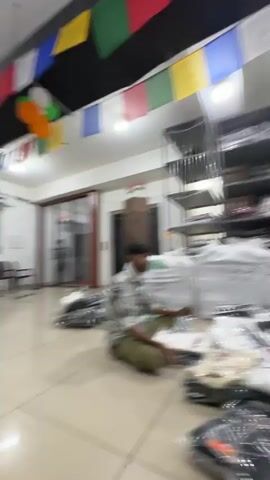 The video shows a well-lit interior space, possibly a warehouse or retail storage area, filled with stacks of clothing. Large white plastic bags, presumably containing more clothing, are prominent throughout the scene. In the foreground, piles of individually packaged t-shirts are neatly arranged. In the background, multiple rows of tall, open shelving units are packed with folded garments. Two men are visible in the middle ground; one is standing and appears to be interacting with a shelf, while the other is crouched down, possibly sorting or examining items. Another person is seated on the floor further back. Prayer flags are strung across the ceiling, adding a decorative element. The overall impression is one of an organized but busy clothing storage and distribution environment.