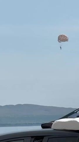 A person is parasailing in a wide-open sky. The parasail is a light brown color and the person is suspended below it. In the distance, there is a body of water with a line of low hills behind it. In the foreground, the top of a black car is visible, with a white suitcase or bag on its roof. The sky is a pale blue and appears to be daytime.