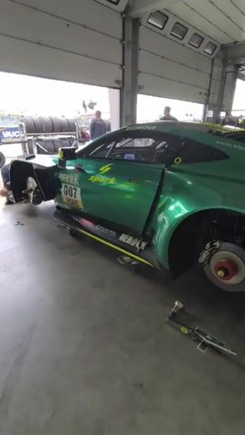 Inside a well-lit garage with a large roll-up door, a green racing car with the number 007 is undergoing maintenance. The car's door is open, and the wheels have been removed, exposing the brake rotors. A torque wrench lies on the concrete floor beside the car. In the background, people are milling around. The car is adorned with sponsor logos. The garage has multiple skylights, and there's another vehicle partially visible behind the main car. The floor is light gray and smooth.