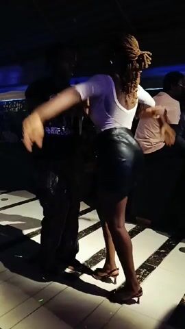 A couple is dancing closely in a dimly lit venue that appears to be a bar or club. A man in dark clothing with colorful patterned pants holds a woman dressed in a white top, a black leather mini-skirt, and high-heeled sandals. She has long dreadlocks. Other people can be seen seated in the background at tables or along a bar. The dance floor has a black and white tiled pattern. The lights overhead shift in color, occasionally displaying blue or green hues.