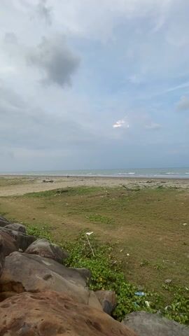 The video shows a cloudy beach scene with the camera panning from right to left, revealing a motorcycle parked in the grassy area. In the background is the ocean, a sandy beach, and some distant figures on the beach.  In the foreground are some large rocks with green vegetation.