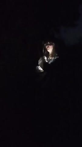 A young person with brown hair, wearing a light-colored shirt and dark pants, is illuminated by a flashlight held in their hand. They are standing in a dark, wooded area, with trees visible in the background. The person appears to be looking at something off-camera. The flashlight beam casts a strong light on their face and upper body, obscuring much of the surrounding environment. The video is shaky, suggesting it was filmed by someone holding the camera while walking.